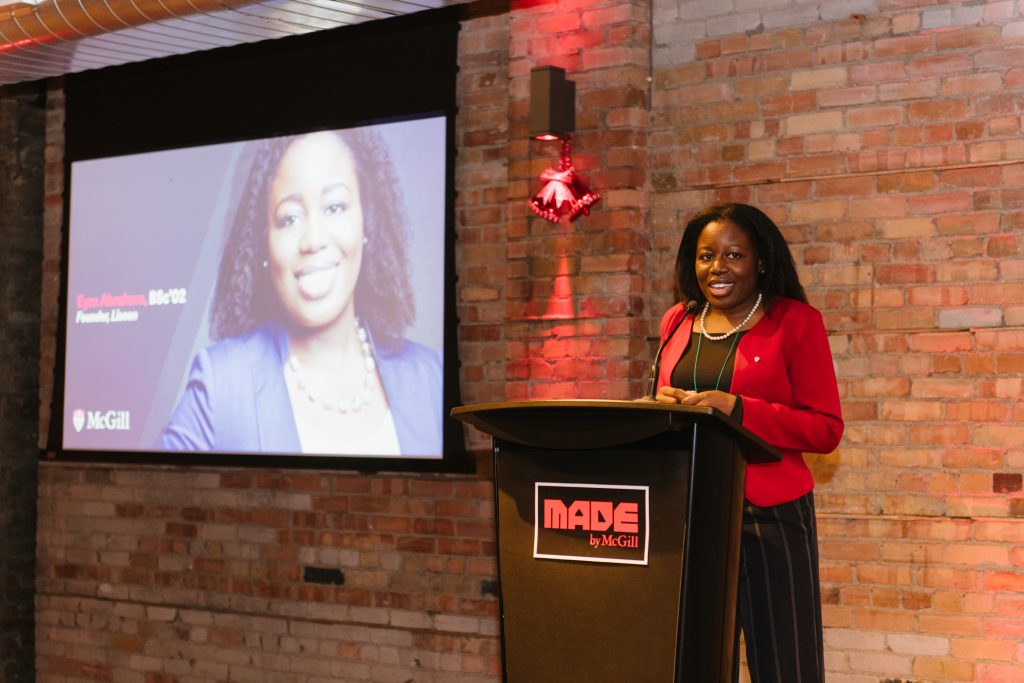 Photo courtesy: McGill University (from Flickr) Eyra on the stage in front of a podium (Made by McGill), brick background will red holiday light and decor. A large screen with Eyra's profile picture to the left can be seen leaning on the brick wall.